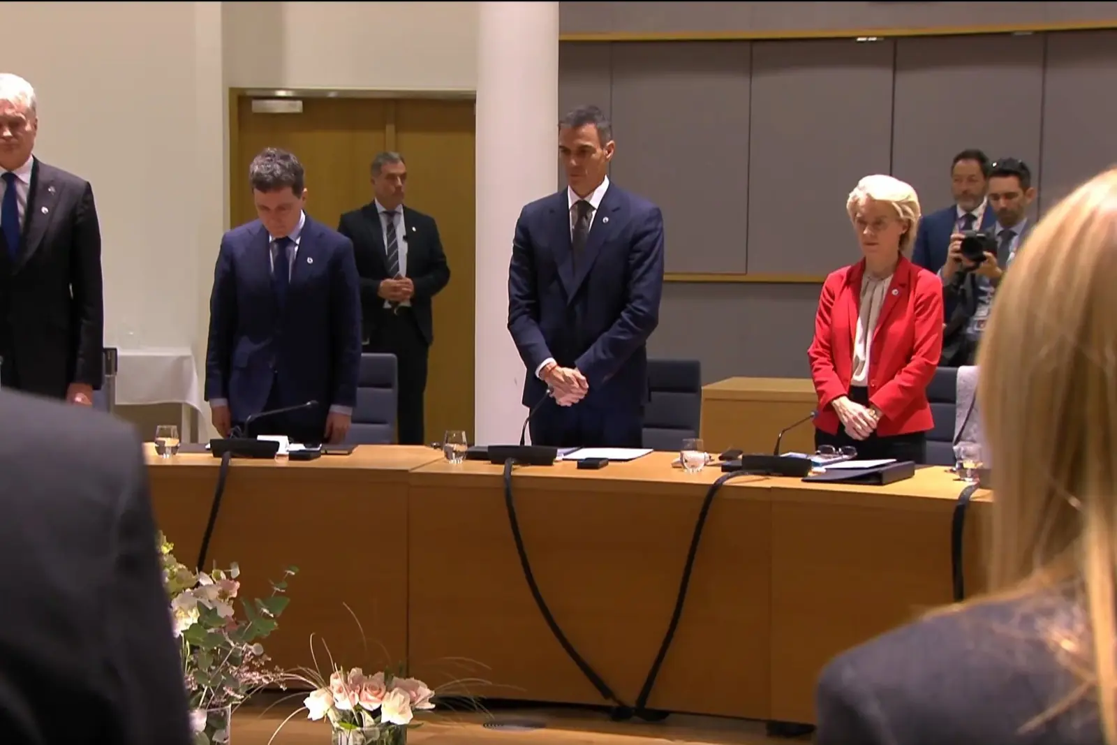 Líderes políticos de pie en una sala de conferencias guardando un minuto de silencio alrededor de una gran mesa de reuniones