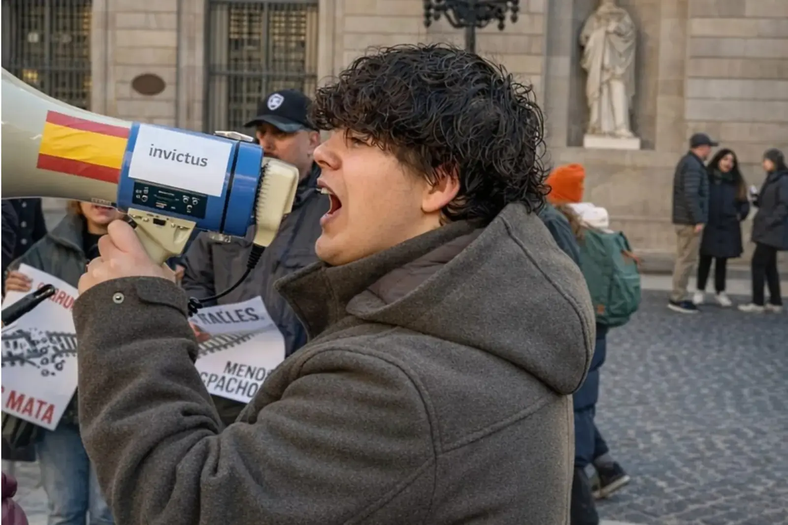 Joven con abrigo gris gritando por un megáfono durante una manifestación en la calle